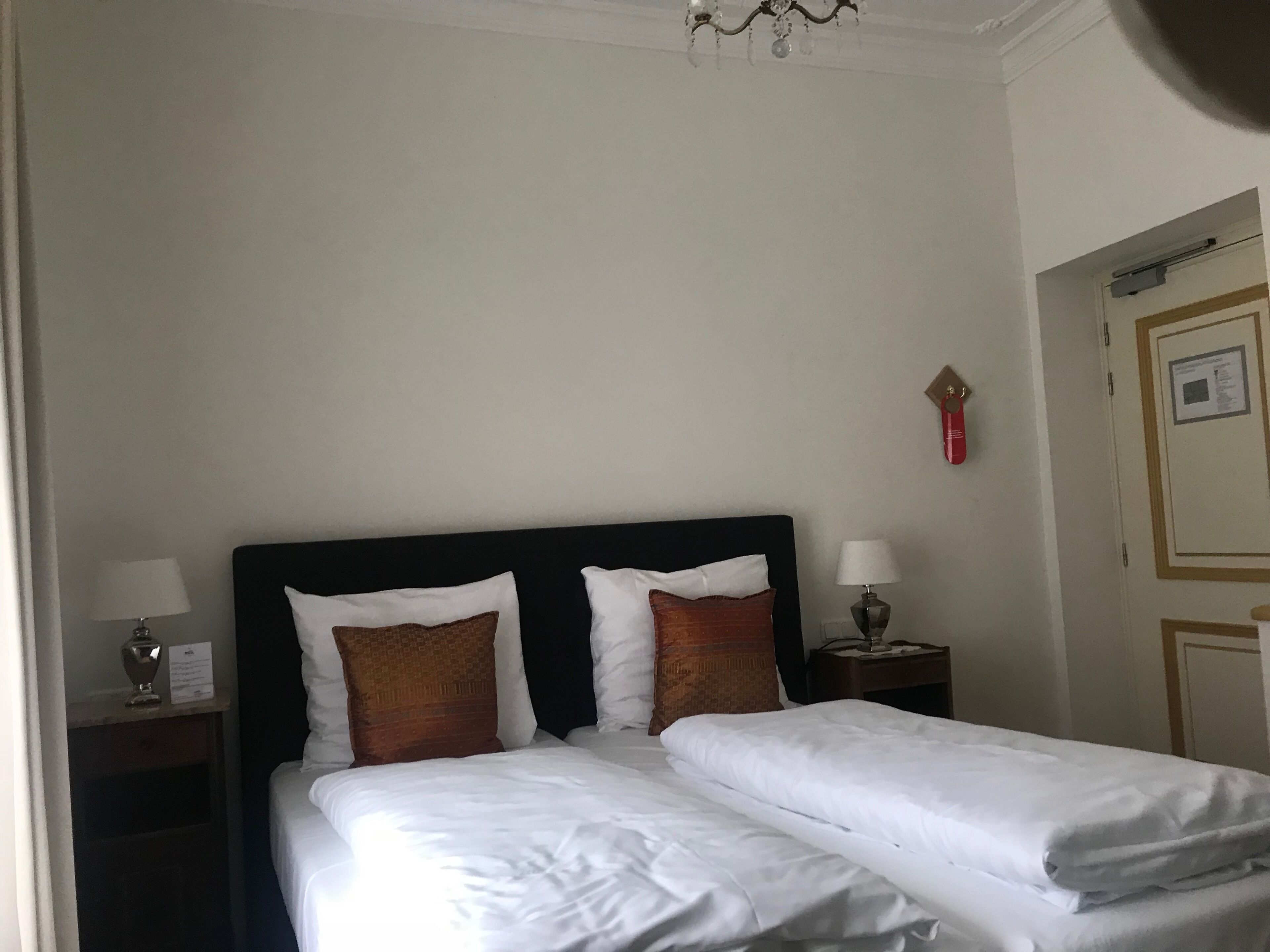 A hotel room with a double bed, two nightstands, a chandelier, and a door with a framed picture.