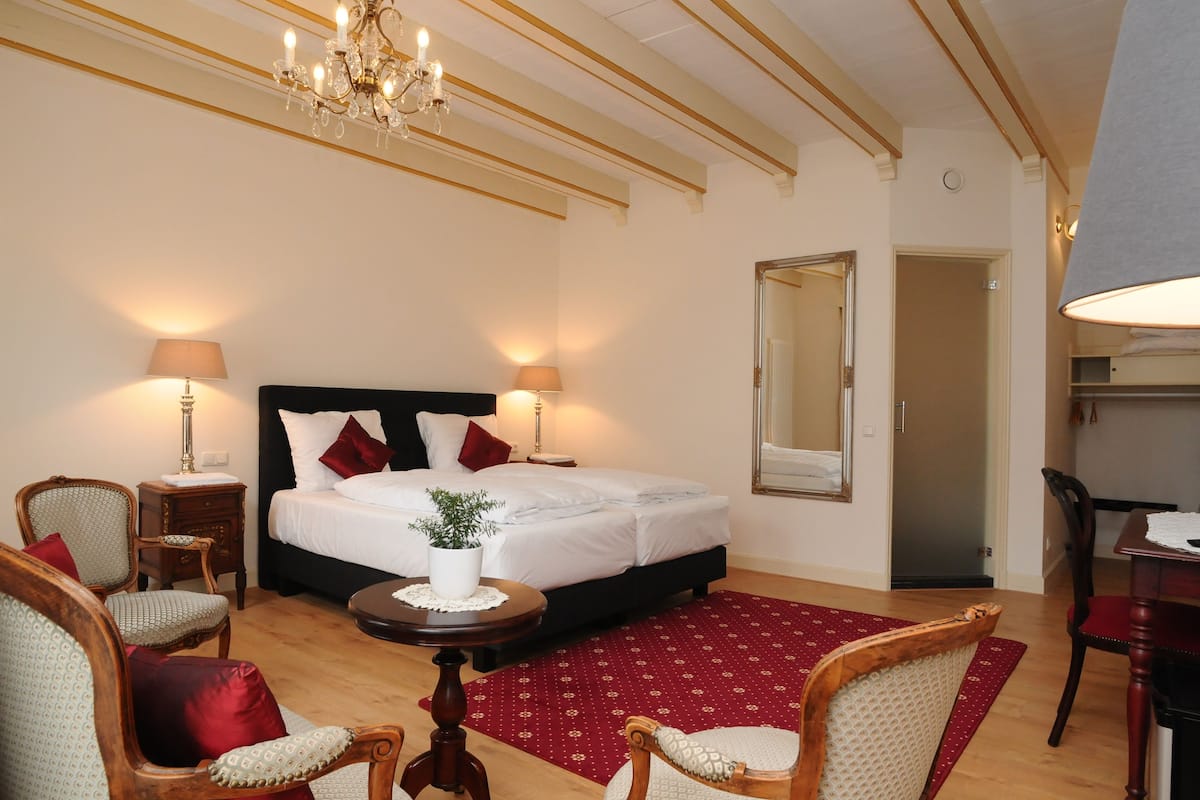 Habitación de hotel con cama, dos mesitas de noche con lámparas, candelabro, espejo, mesa de comedor y alfombra roja estampada.