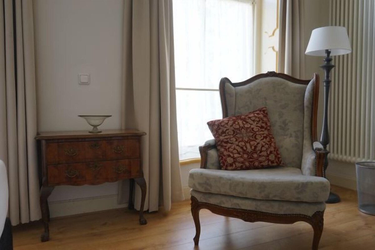 A room with a wooden floor, a patterned armchair with a cushion, a small wooden side table with a bowl, and a lamp.