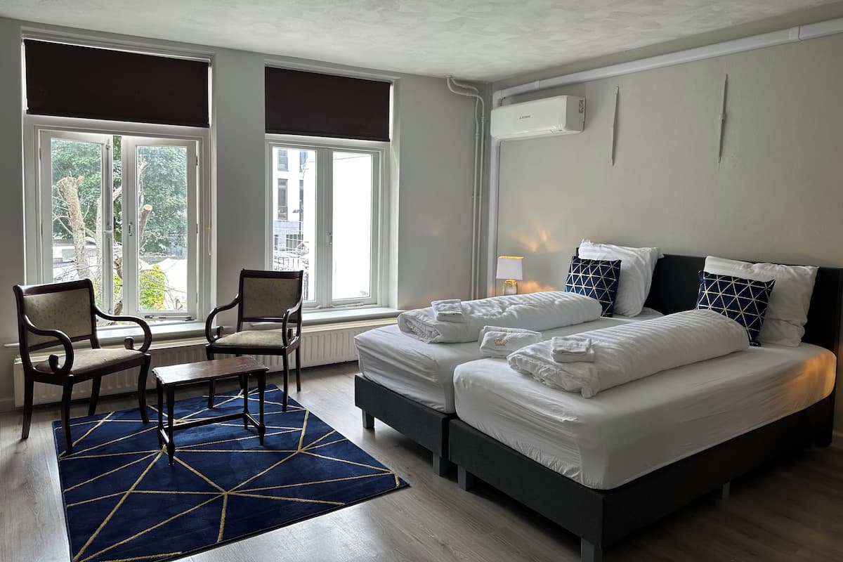Habitación de hotel con dos camas, una silla, una mesa y un ventanal con persianas.