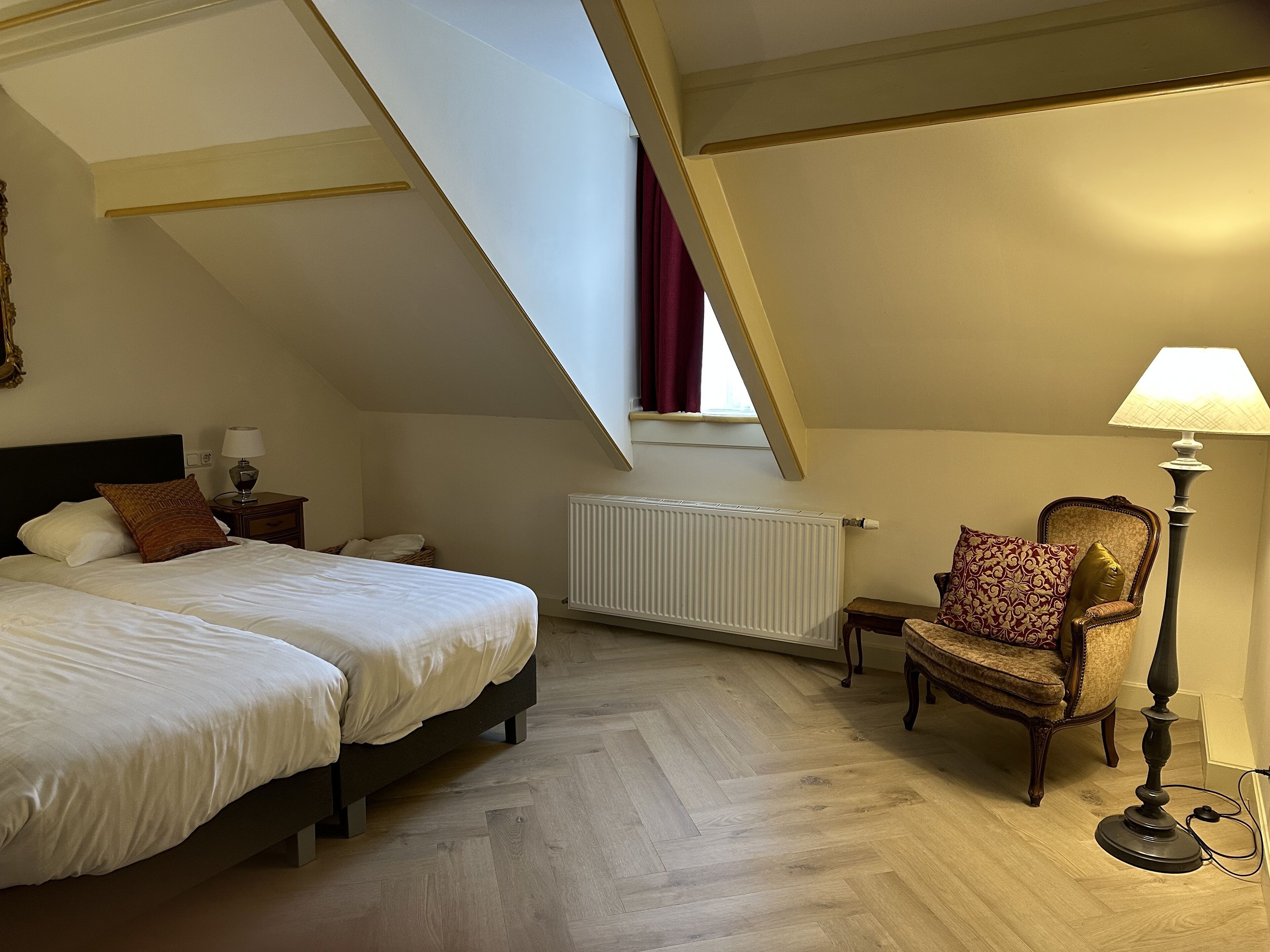 Een slaapkamer met een bed, een stoel, een lamp, een radiator en een raam met gordijnen.