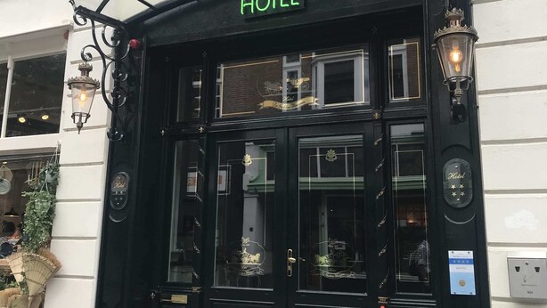 Een hotelentree met een zwart gevel, een 'HOTEL'-bord boven de deuren, en vlaggen van Nederland, België en de Europese Unie.