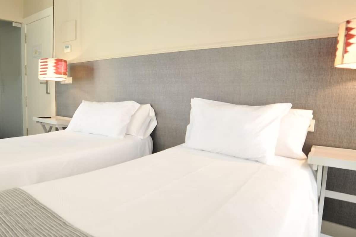 Habitación de hotel con dos camas, cabecera gris y una lámpara roja a la izquierda.