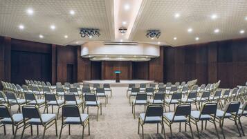 Un salón de conferencias con filas de sillas mirando hacia un escenario, una alfombra estampada y un diseño moderno en el techo.