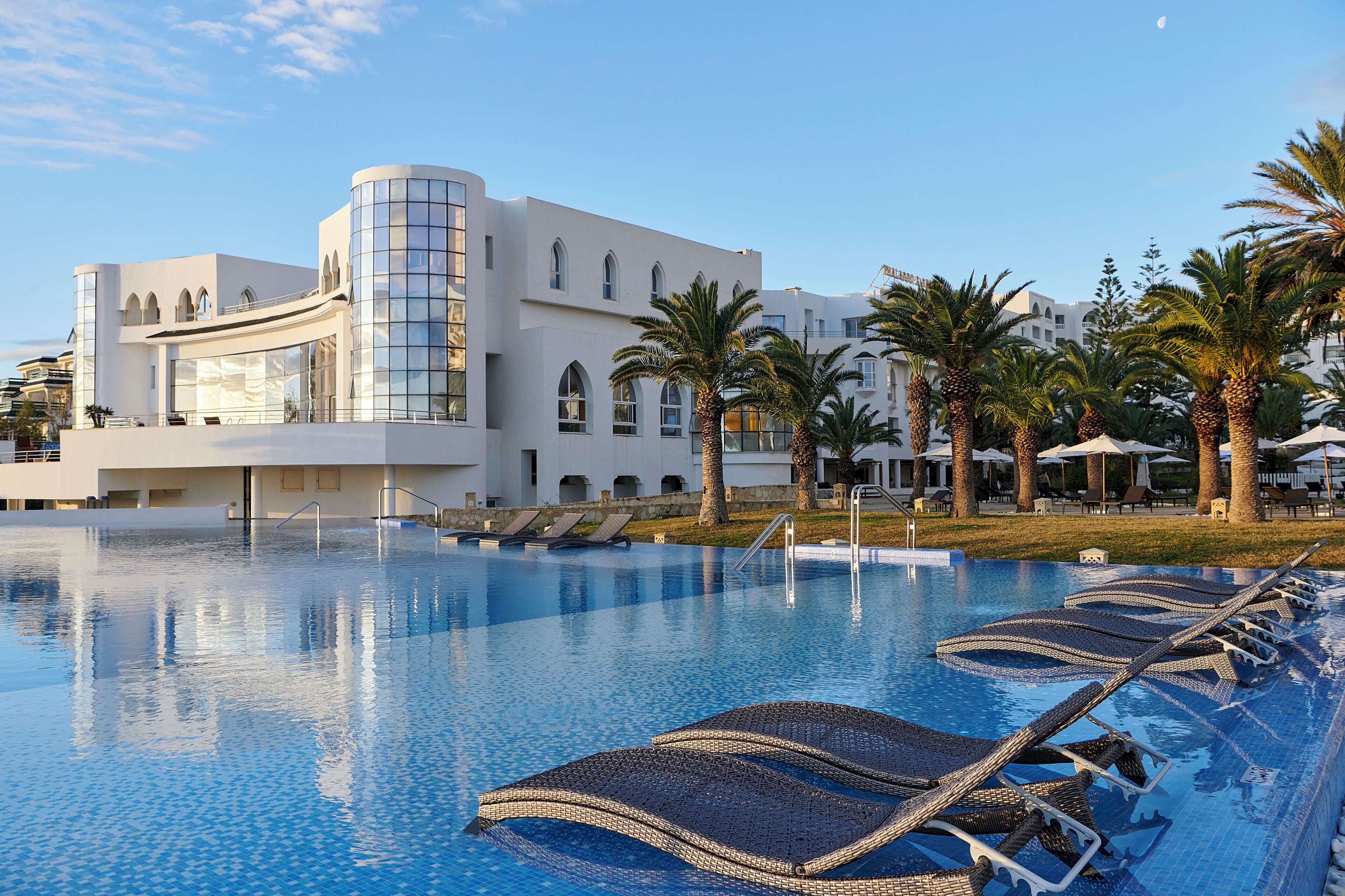 A modern hotel with a swimming pool, palm trees, and lounge chairs.