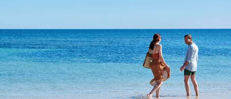A couple walking on a beach with clear blue water and a sandy shore.