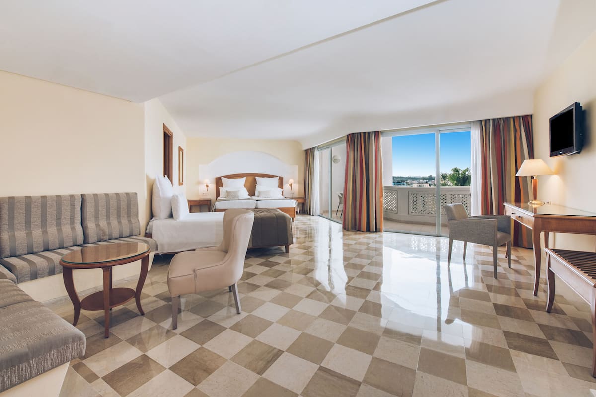 Habitación de hotel con piso a cuadros, un sofá, cama con cabecera, escritorio, una silla y televisor en la pared.