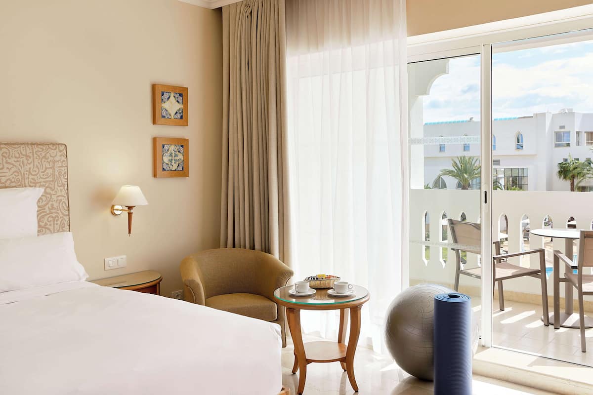 A hotel room with a bed, a chair, a small table with cups and a tray, a yoga ball, and a balcony with chairs and a table.