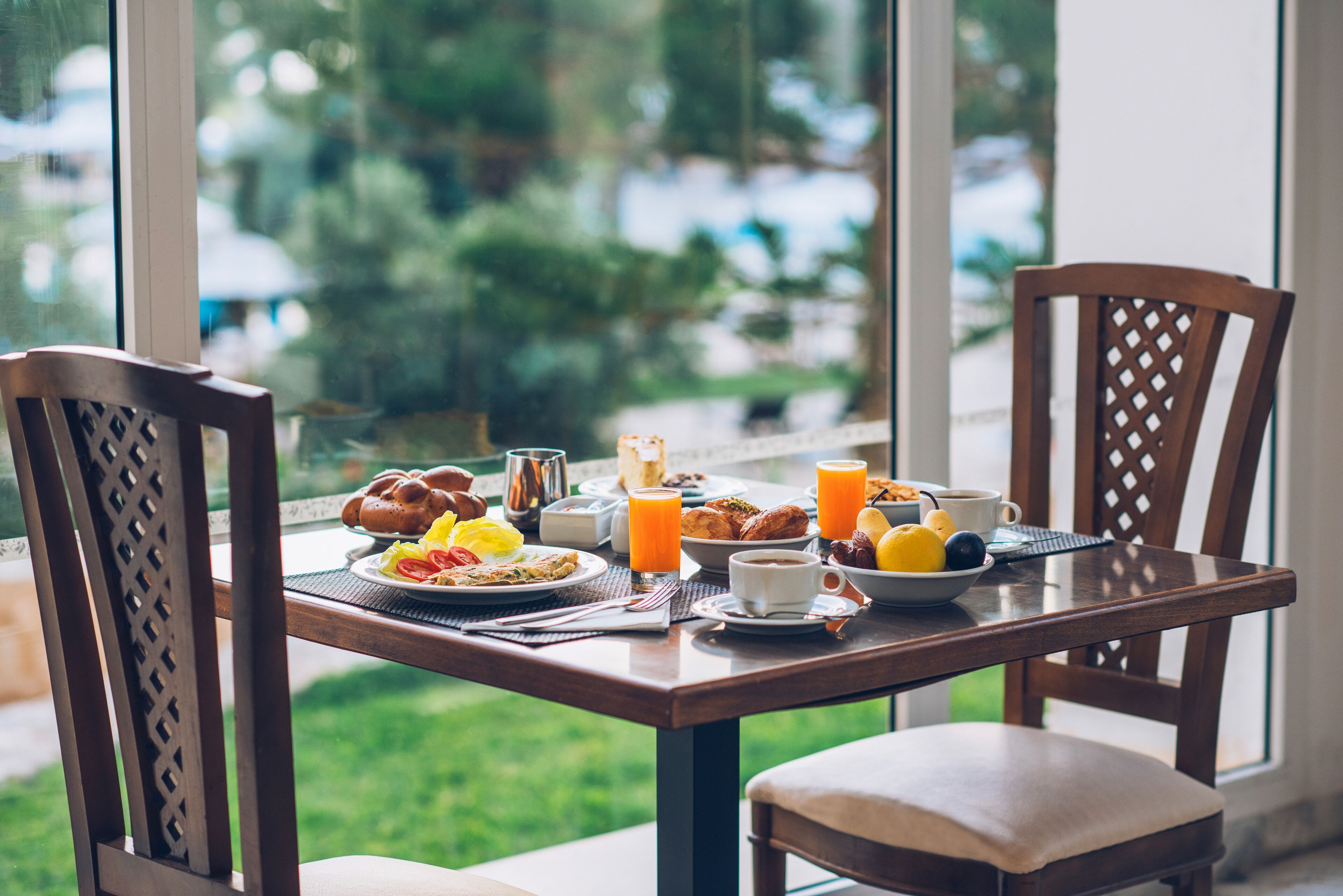 A breakfast table with various dishes and drinks, set against a window with a view of greenery.