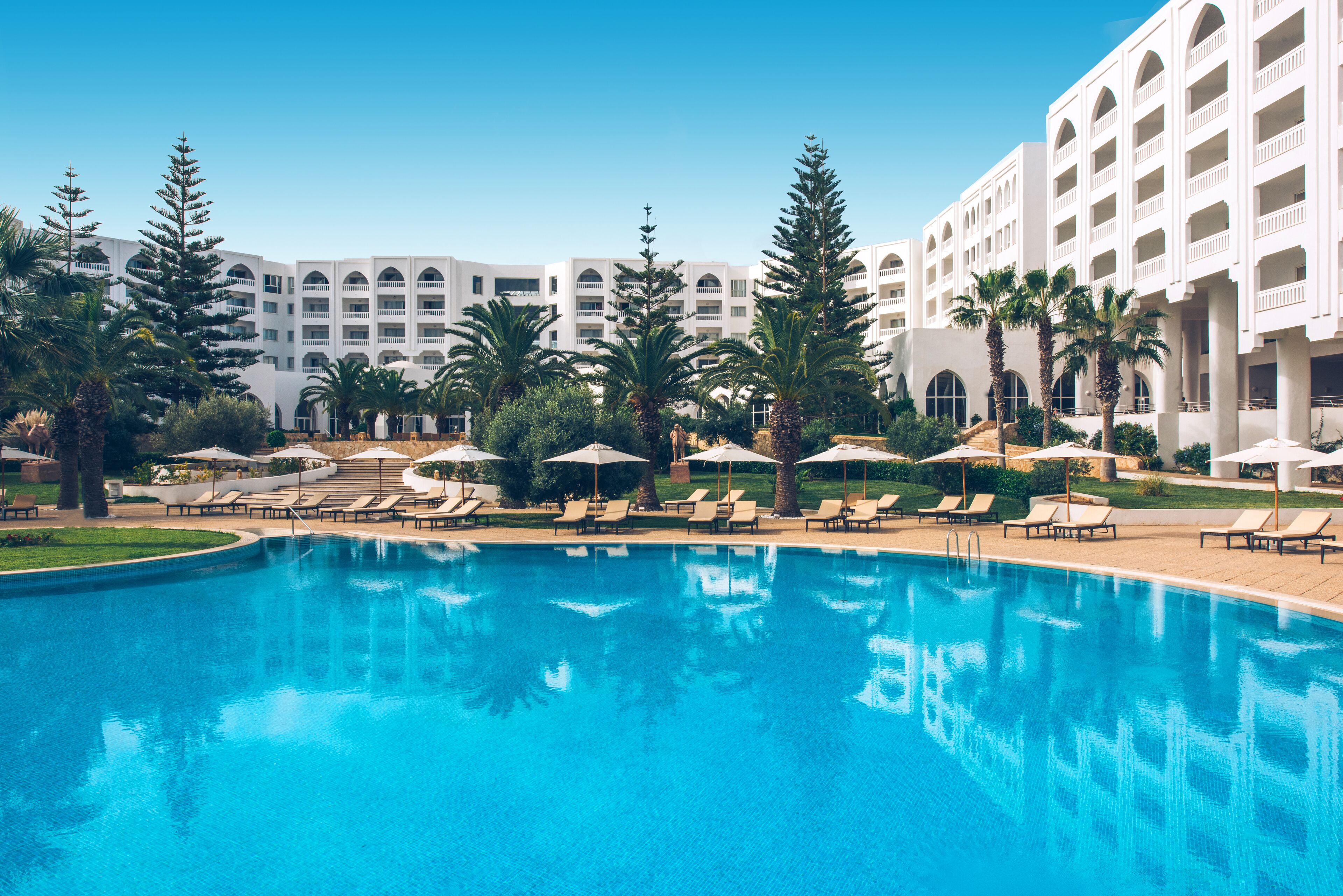 Una piscina con sillas de descanso y sombrillas, rodeada de palmeras y un edificio hotelero con varios balcones.
