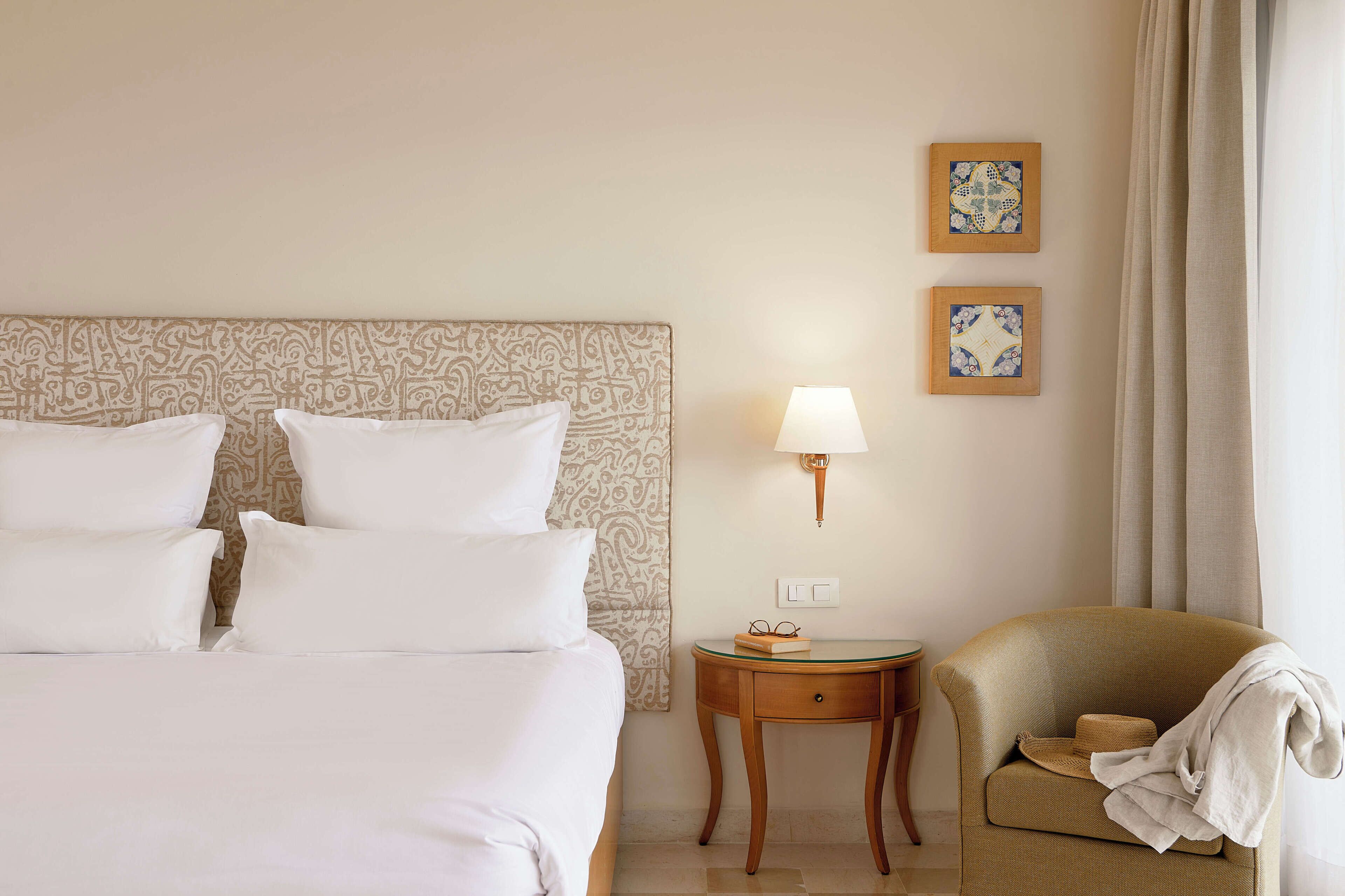A hotel room with a large bed, a bedside table with a lamp, a chair, and two framed pictures on the wall.