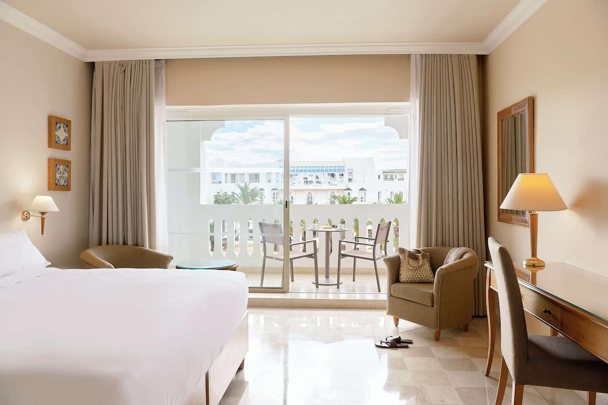 A hotel room with a bed, a desk, a chair, a table, and a balcony with a view of buildings.