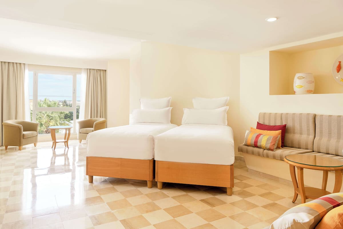 Una habitación de hotel con piso a cuadros, una cama con dos almohadas, un sofá con cojines, una pequeña mesa redonda y una ventana con cortinas.