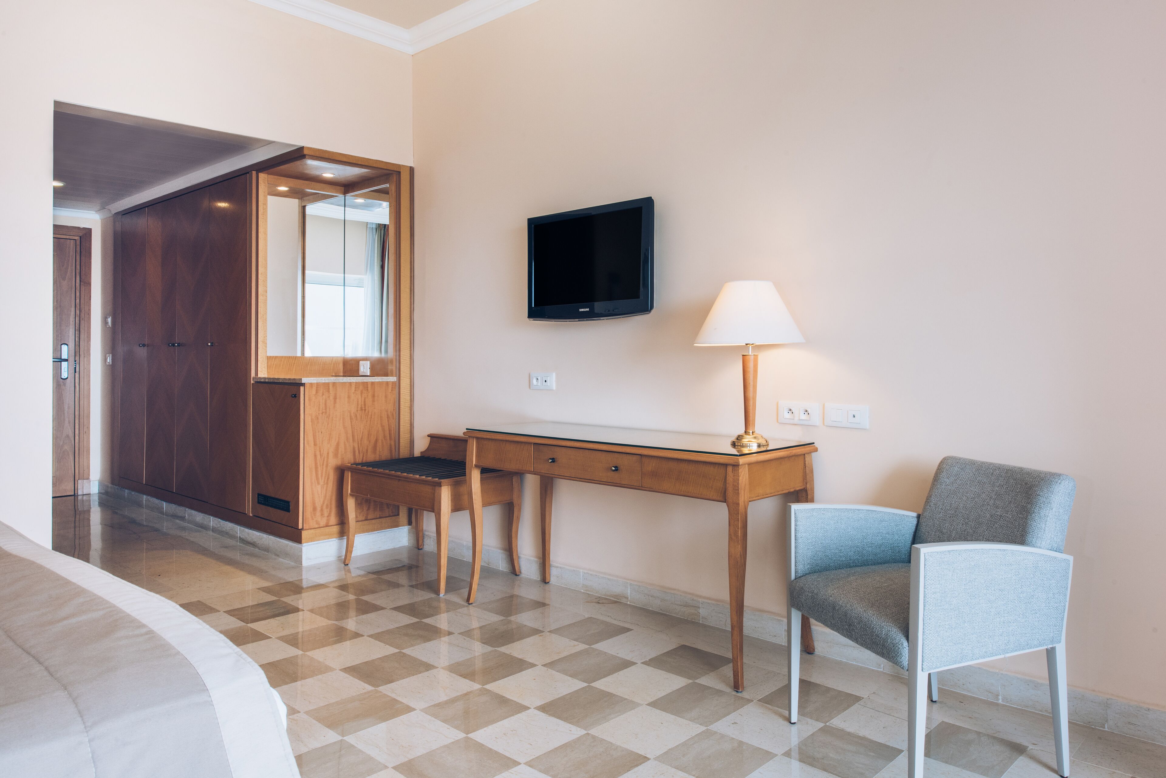Quarto de hotel com chão xadrez, uma mesa com cadeira, uma televisão fixada na parede e um abajur.