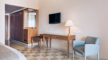 Quarto de hotel com chão xadrez, uma mesa com cadeira, uma televisão fixada na parede e um abajur.