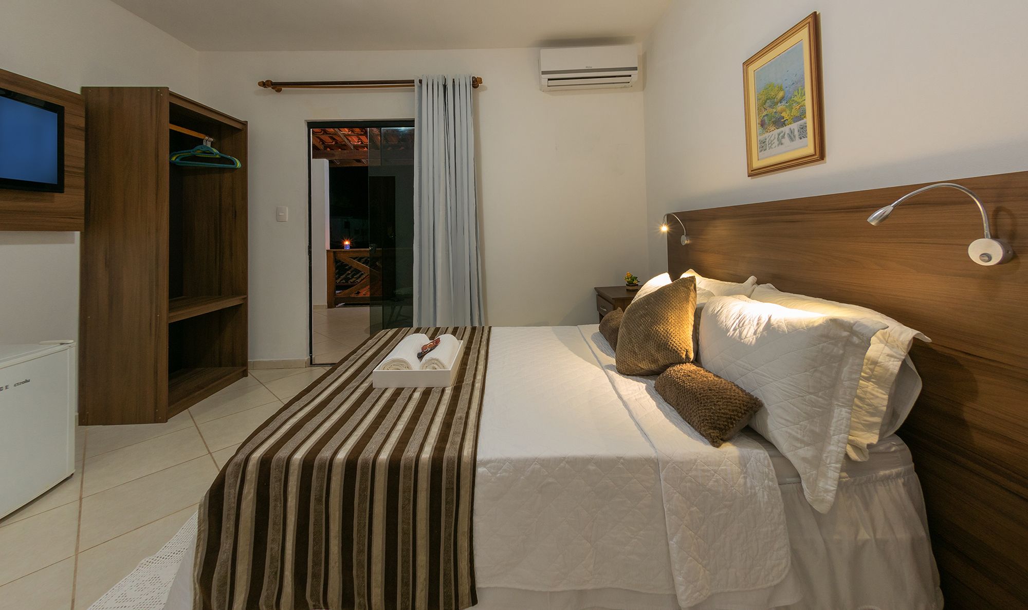 Una habitación de hotel con una cama, un armario de madera, un televisor, un cuadro y un equipo de aire acondicionado.