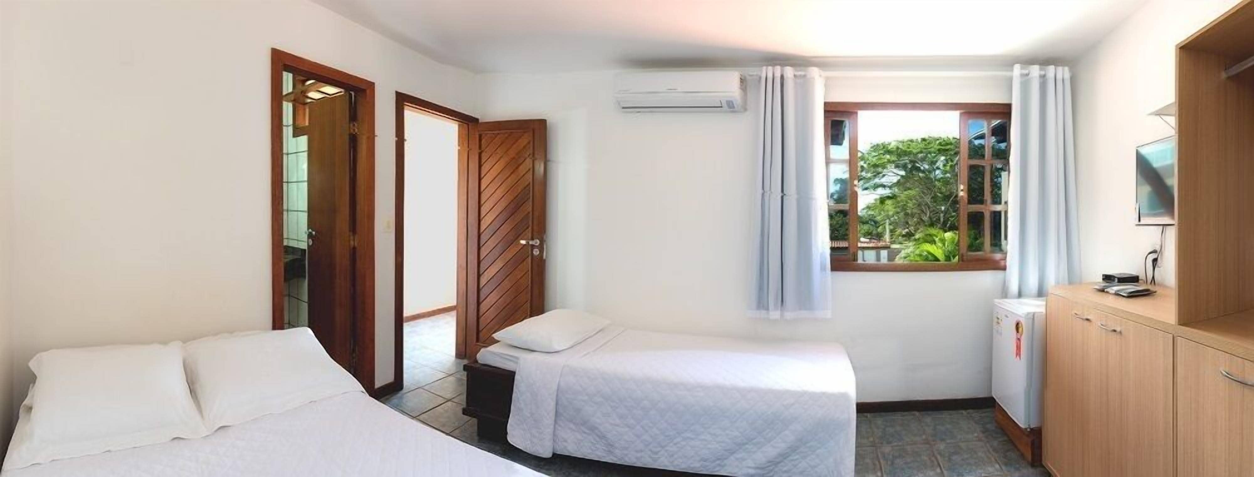 A room with two single beds, a small kitchenette, a window with curtains, and an air conditioning unit.