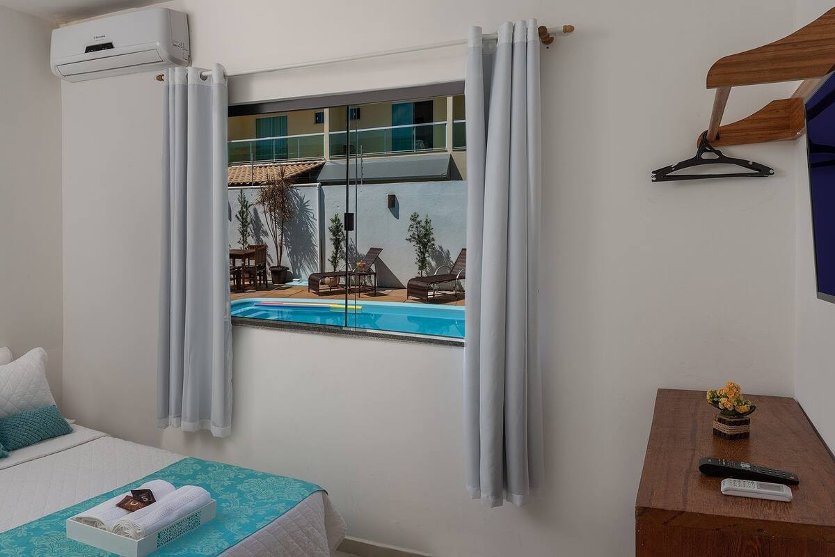 A hotel room with a bed, a window with curtains, a small table with a remote control, and a TV on the wall.