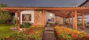 Un espacio al aire libre cubierto con un muro de piedra, una pérgola de madera y un camino que conduce a la entrada de un edificio.