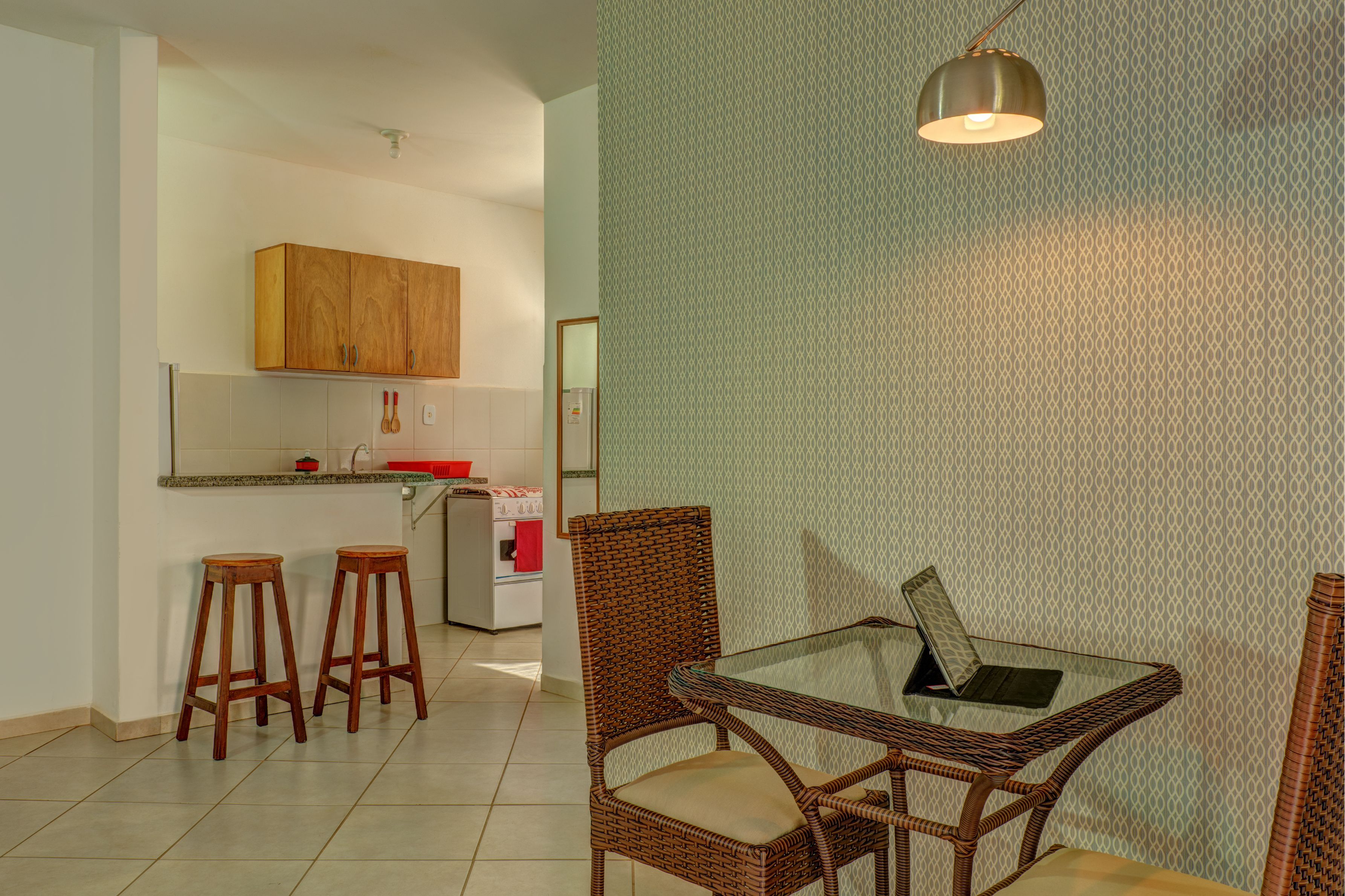 A kitchen with wooden cabinets, a refrigerator, and a small countertop. There are two wooden stools and a wicker chair around a glass table with a laptop.