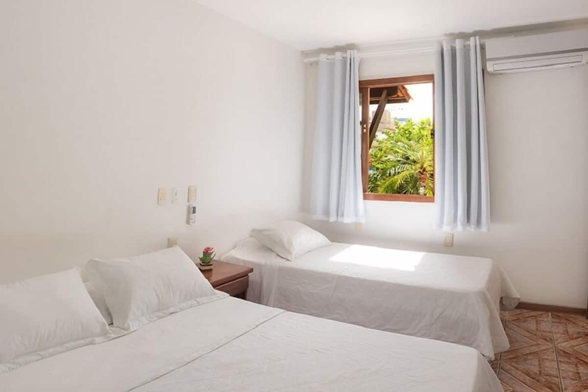 A room with two single beds, a window with curtains, a nightstand, and a view of greenery outside.