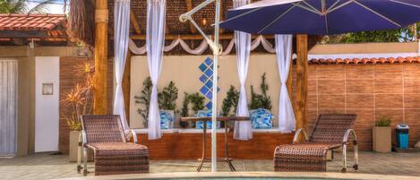 Un área de piscina con dos sillones de mimbre, una mesa con mantel blanco y un gran paraguas.