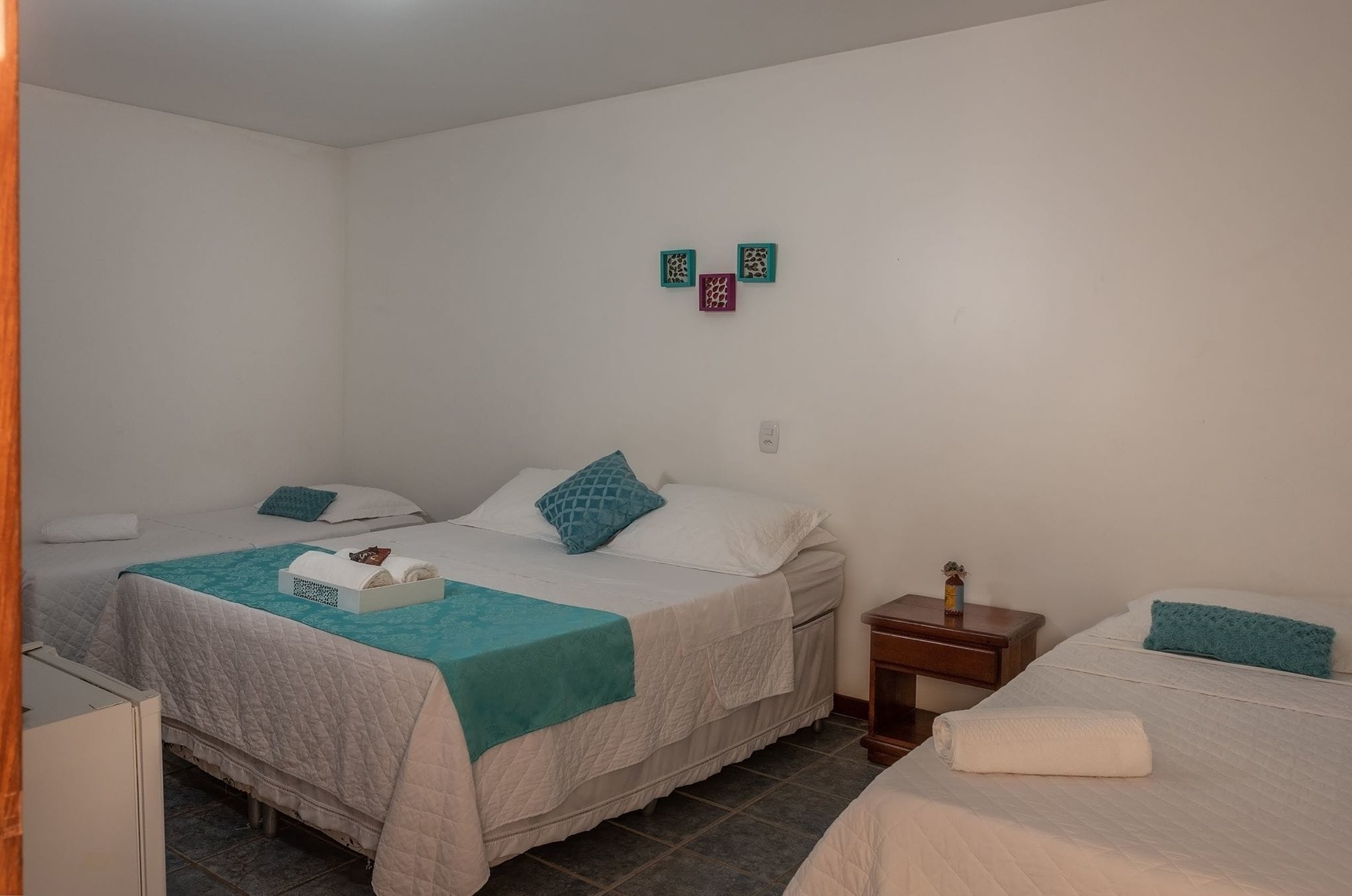 A hotel room with two beds, each with a white and teal bedspread, and a small bedside table with a lamp.
