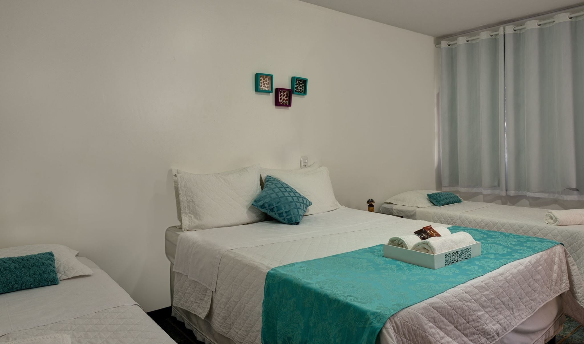 Habitación de hotel con dos camas, cada una con cubrecama blanco y almohadas turquesa. Hay una mesita con mantel turquesa y una caja de artículos de aseo.