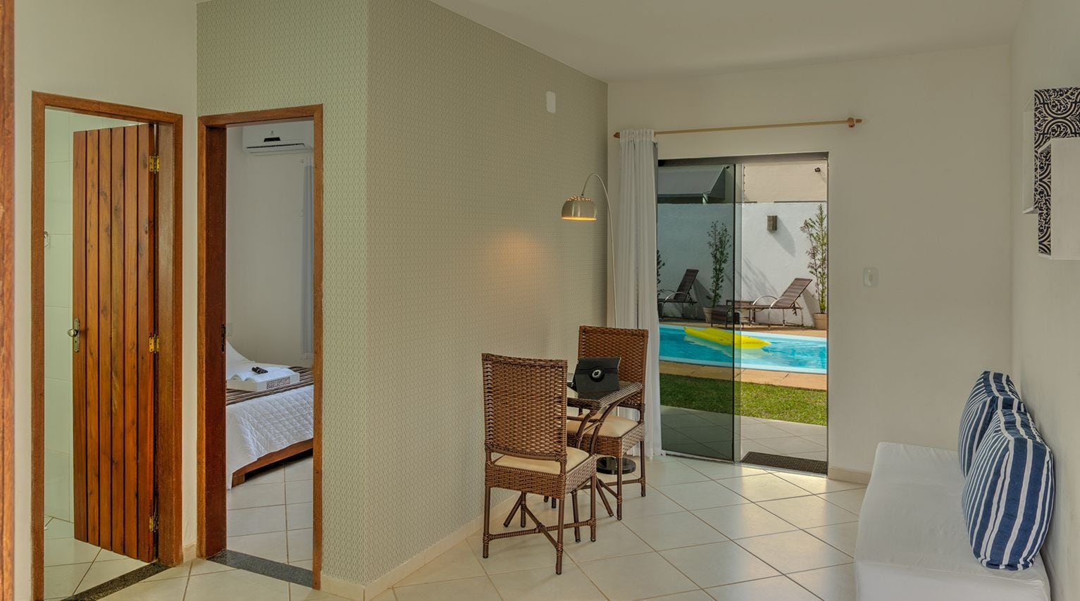 Habitación de hotel con cama, zona de comedor con sillas de mimbre, vista a una piscina y puerta corrediza de vidrio que da a un área exterior.