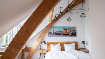A bedroom with a sloped ceiling, wooden beams, a bed with white linens, two bedside tables with lamps, and a painting of a coastal scene.