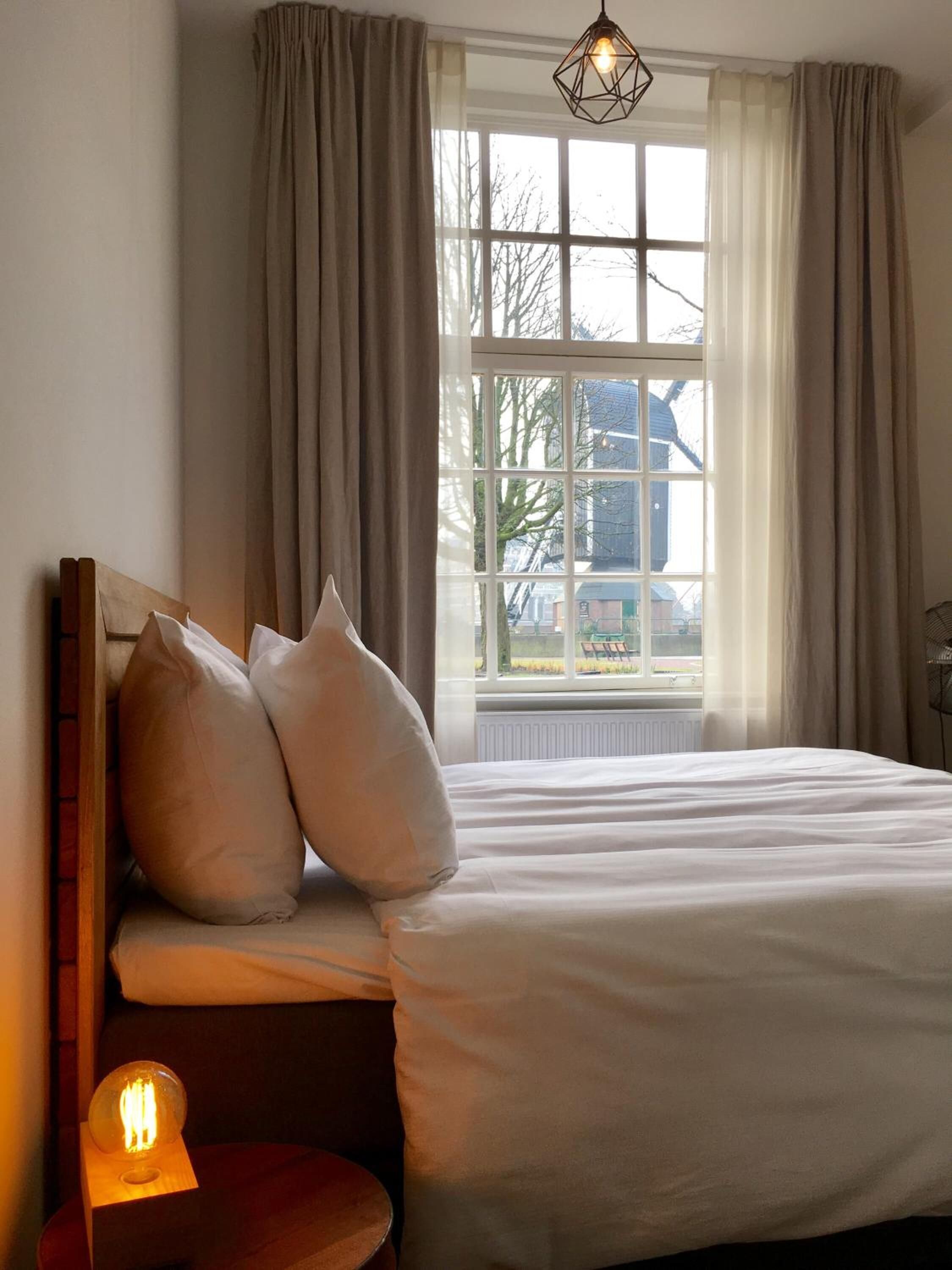 Ein Schlafzimmer mit einem großen Bett, einem Nachttisch mit Lampe und einem Fenster mit Vorhängen.
