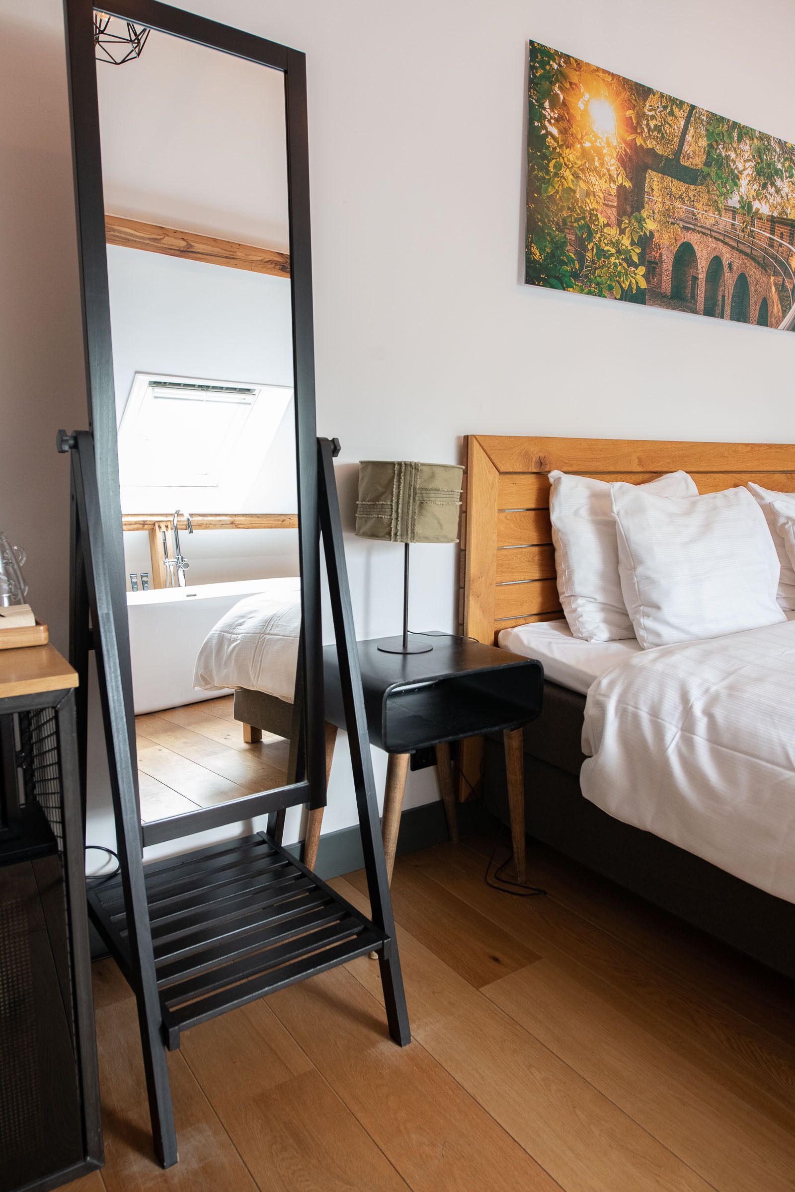 A bedroom with a wooden headboard, a bed with white bedding, a nightstand with a lamp, and a large mirror.