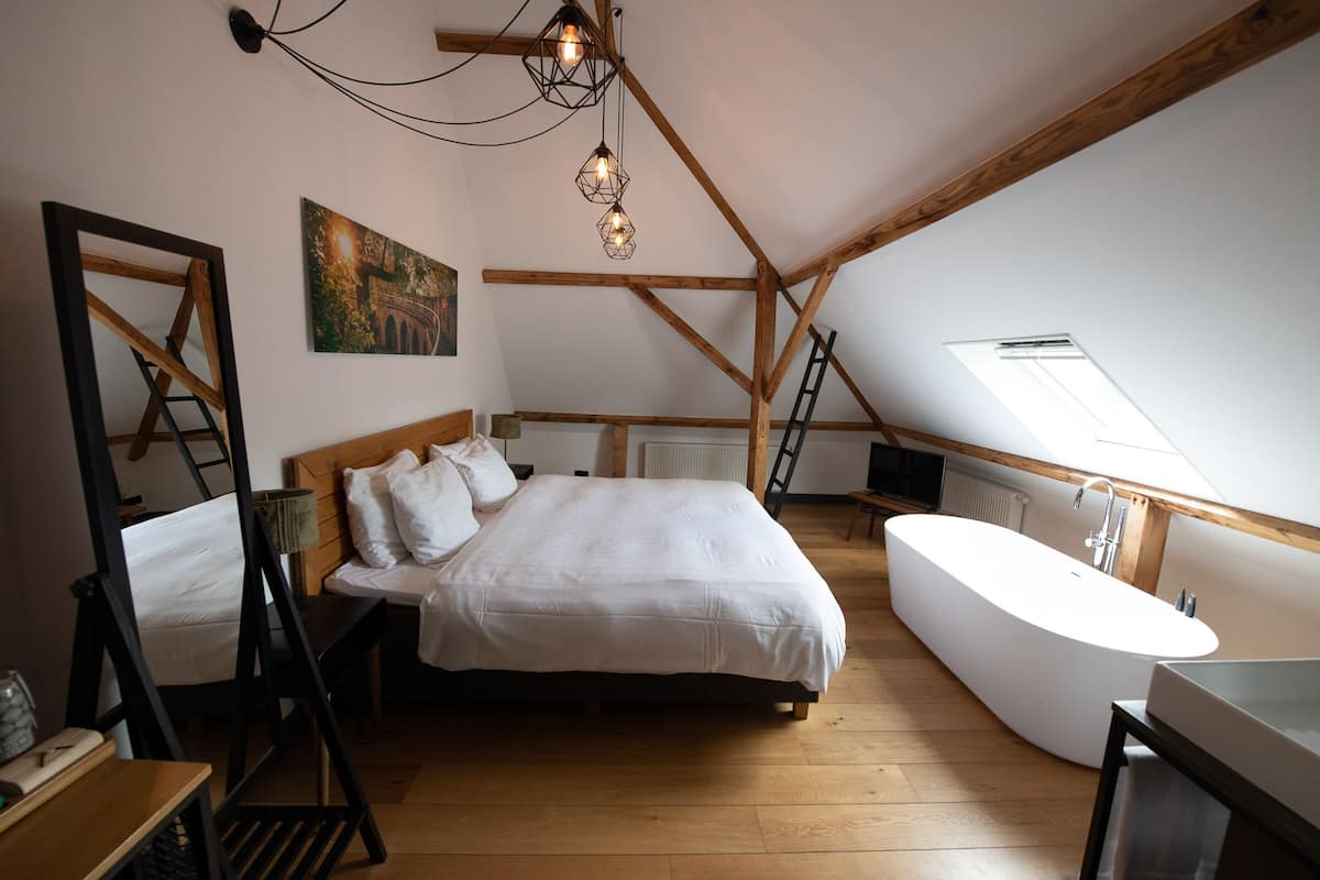 Een slaapkamer met een groot bed, een witte badkuip, houten balken, een dakraam en een schilderij aan de muur.