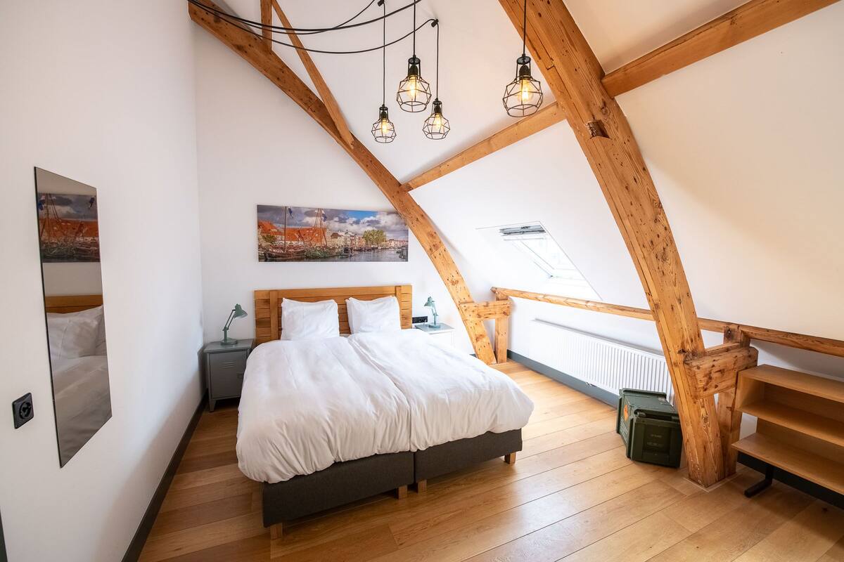 Een slaapkamer met een houten plafond, een bed, een nachtkastje, een lamp en een schilderij aan de muur.
