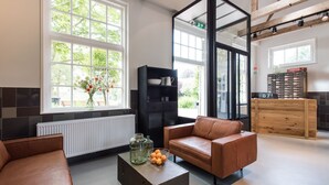 Ein modernes Wohnzimmer mit einem braunen Ledersofa, einem schwarzen Bücherregal, einem Couchtisch mit Dekoration und großen Fenstern mit Blick auf Grünflächen.