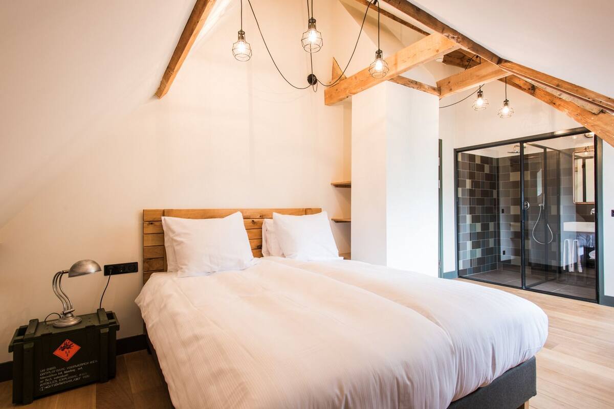 Een slaapkamer met een houten plafond, een bed met wit beddengoed, een bedlampje en een douchecabine van glas.