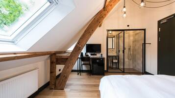 A bedroom with a skylight, wooden beams, a bed, a desk with a computer, and a shower room.