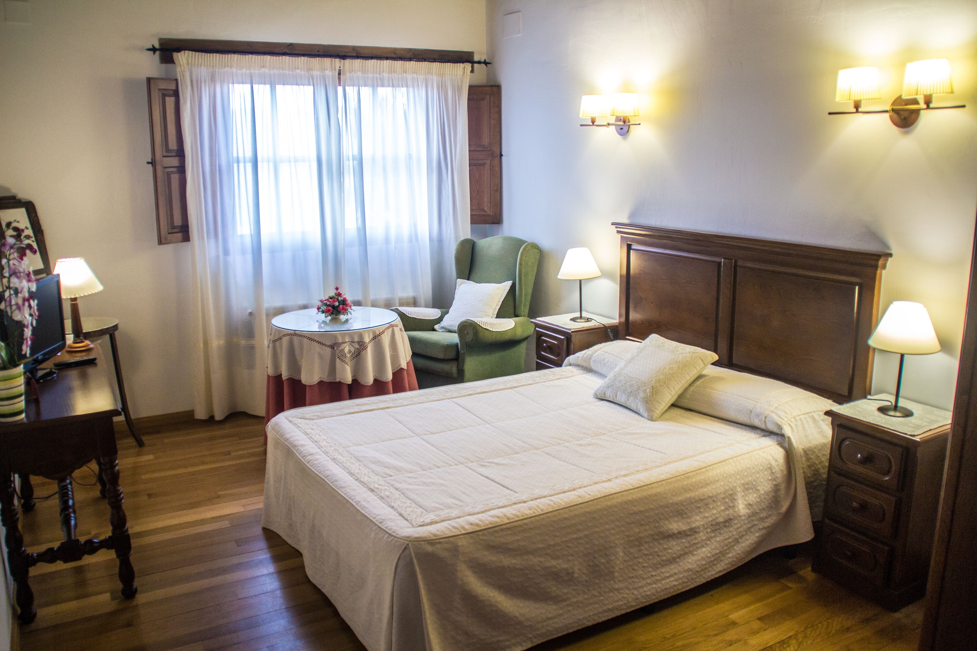 A neatly arranged bedroom with a large bed, a small round table, a green armchair, and two bedside lamps.