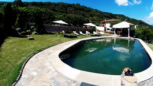 Una piscina con baldosas de piedra, rodeada de césped y un muro de piedra. Hay sillas de descanso y sombrillas.