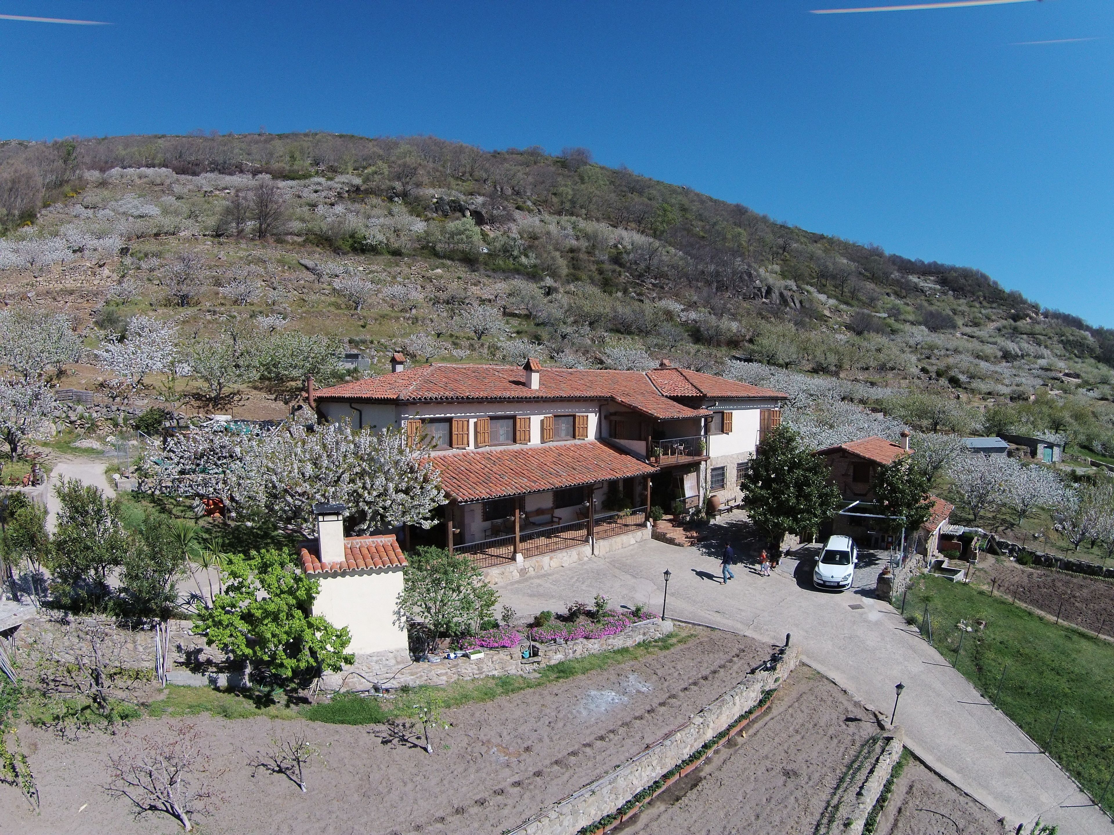 Una casa de dos pisos con techo de tejas, rodeada de un jardín y un paisaje montañoso.