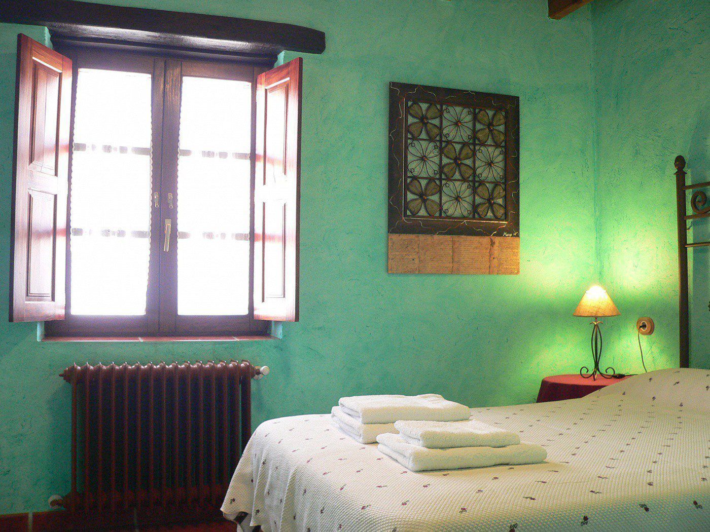 A bedroom with a bed, a radiator, a window with open shutters, and a wall-mounted lamp.