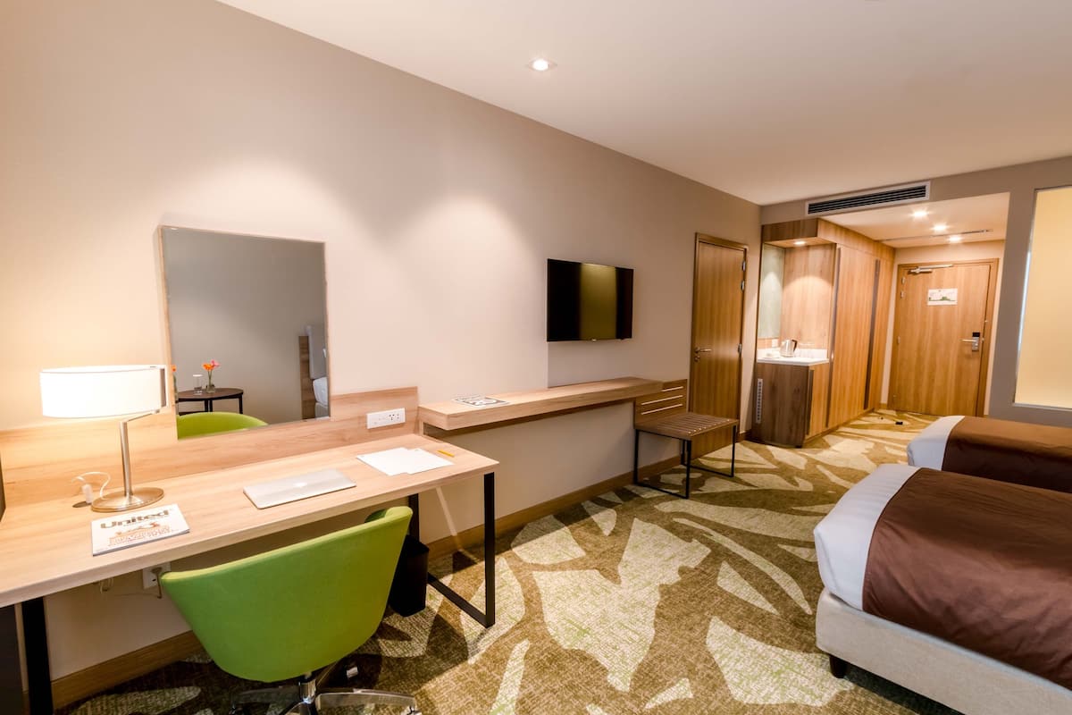 A hotel room with a desk, a green chair, a bed, a TV, and a mirror.