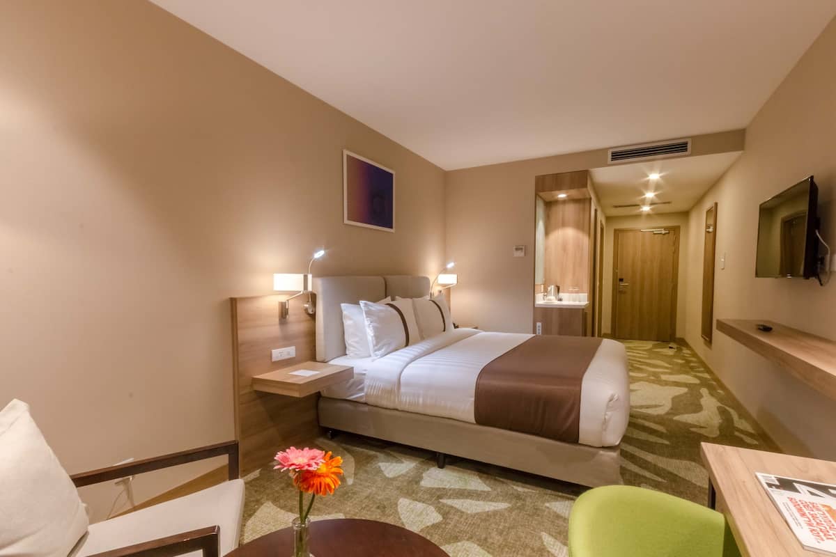 A hotel room with a bed, bedside table, lamp, chair, and a small table with a vase of flowers.