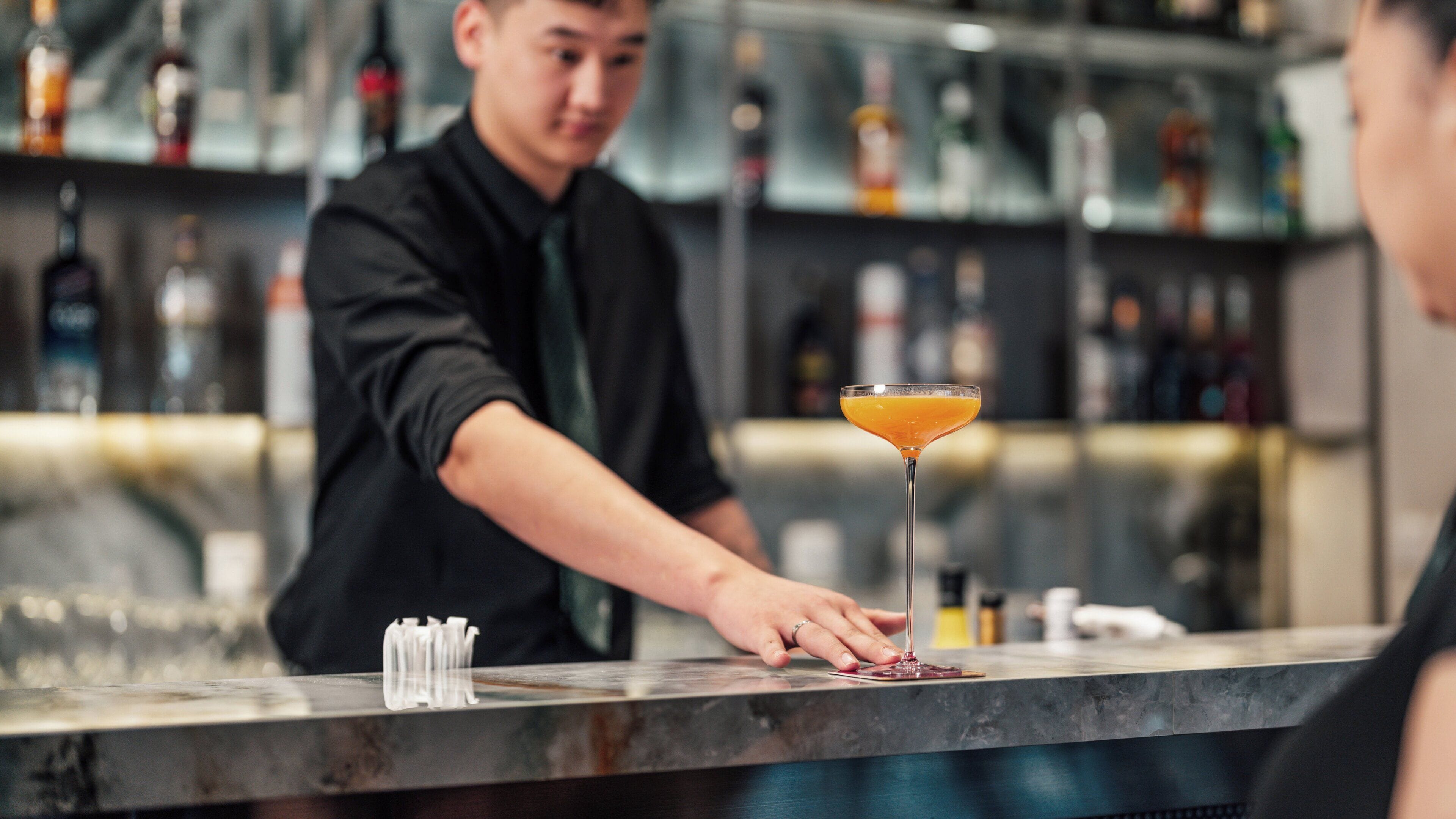 A bartender preparing a drink at a bar with a variety of bottles in the background.
