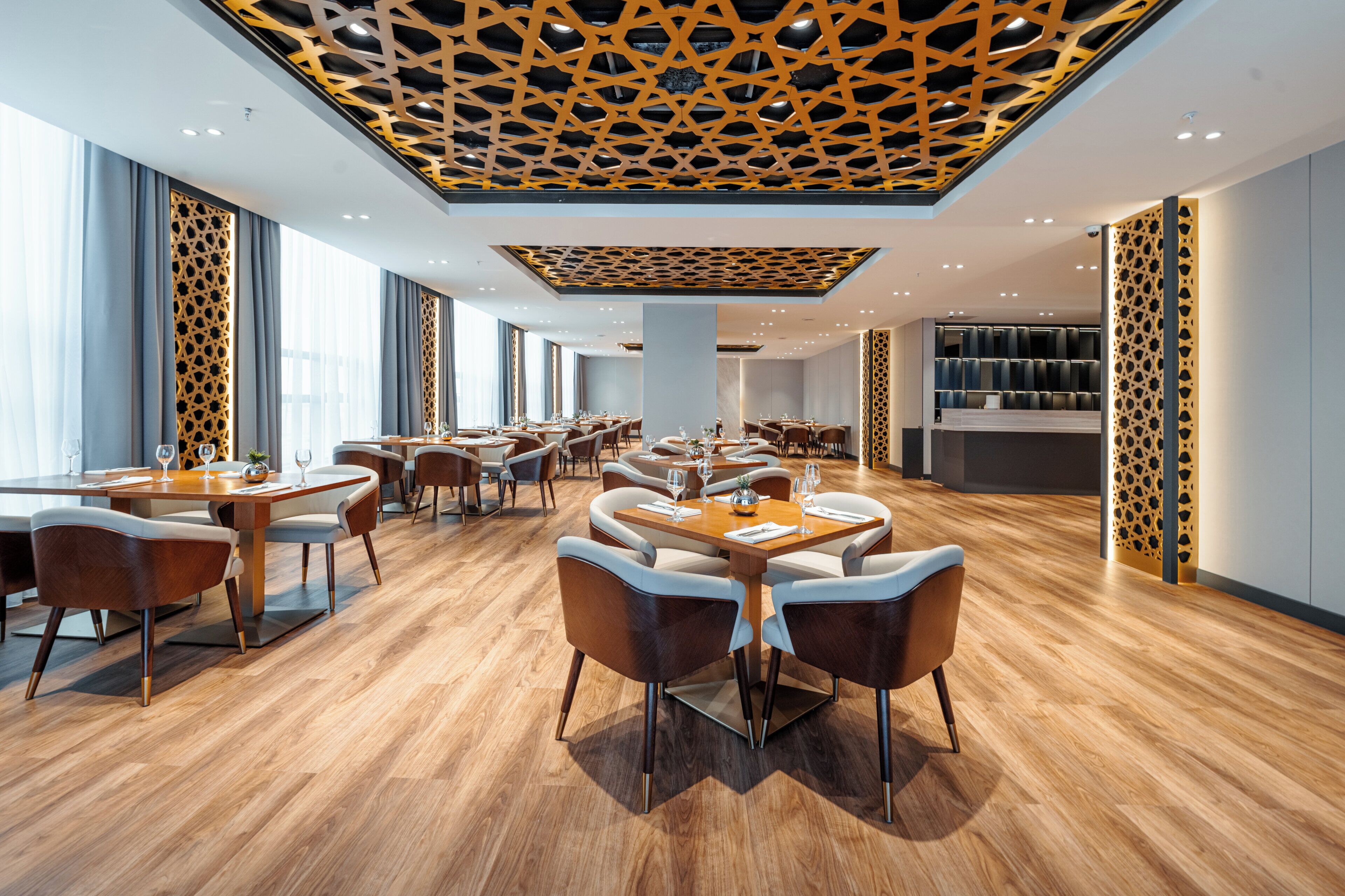 A modern restaurant with wooden flooring, multiple dining tables, and a patterned ceiling.