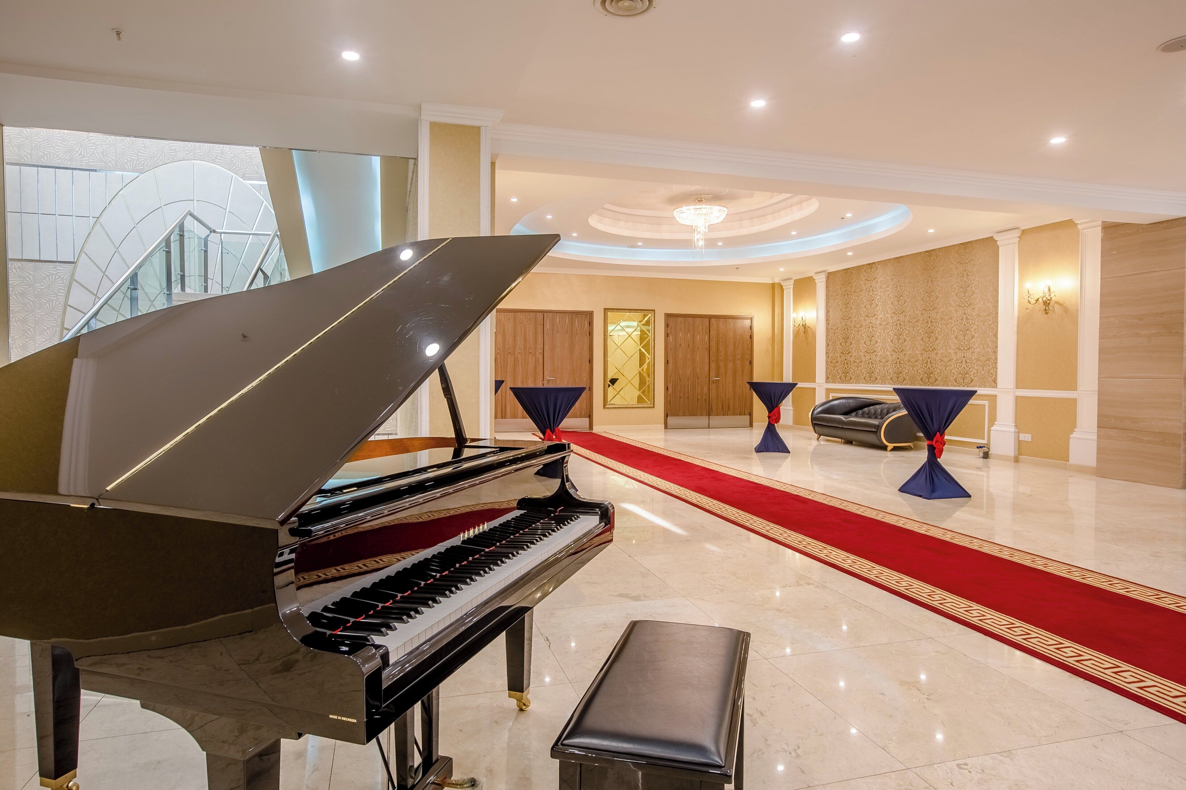 Um piano de cauda em um hall amplo com carpete vermelho, um candelabro e uma área de estar com sofá e poltronas.