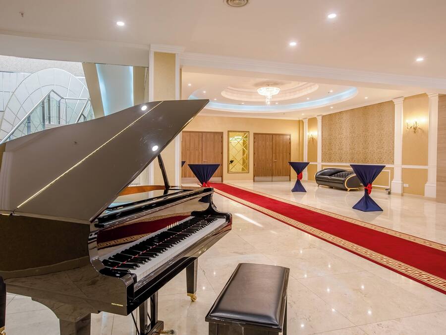Un magnifique piano à queue trône dans un vaste hall d’entrée, agrémenté d’un tapis rouge, d’un lustre et d’un espace salon avec un canapé et des fauteuils.