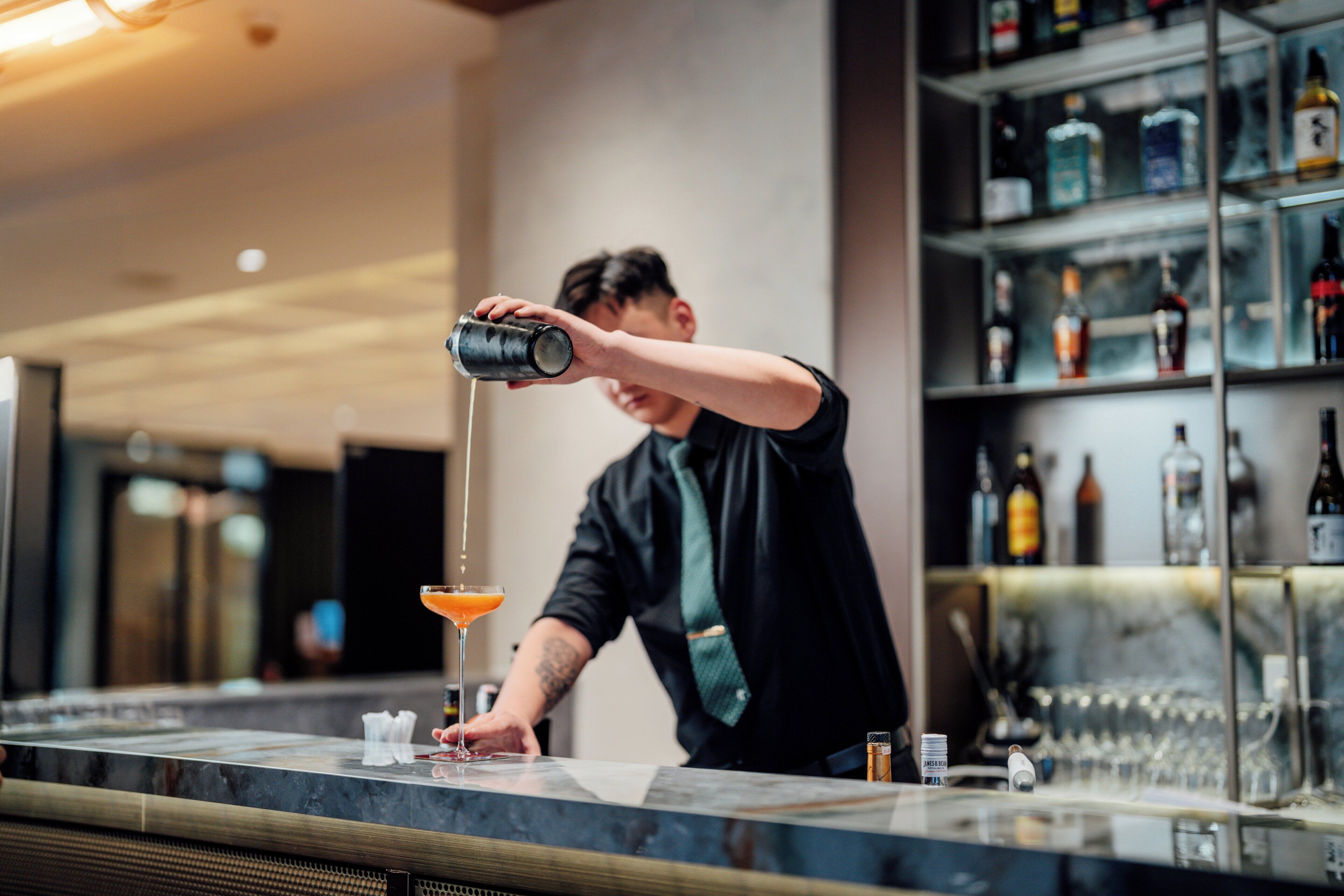 Un barista versa un cocktail in un bicchiere da martini dietro un bancone con scaffali pieni di bottiglie.
