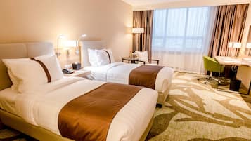 Two large beds with white linens and brown accents, a desk, chair, and table with a vase of flowers, and a lamp.