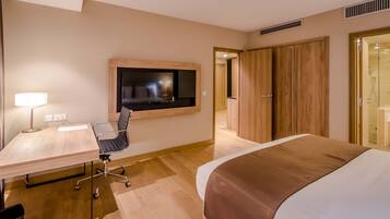 A hotel room with a bed, a desk with a chair, a TV mounted on the wall, and a wooden wardrobe.